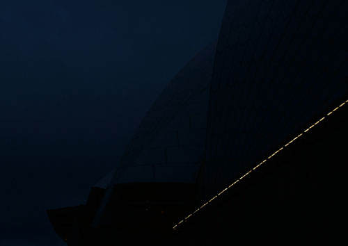 A low contrast night photo of the Sydney Opera House where you are led into a landscape of darkly silhouetted shells by a trail of lights against a deep navy sky.