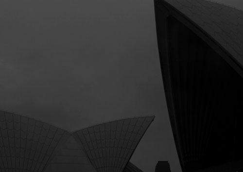 A low contrast night photo of the Sydney Opera House in black and white with a large fraction of a shell dominating the front of the photo and a smaller silhouette of a double shell in the background.