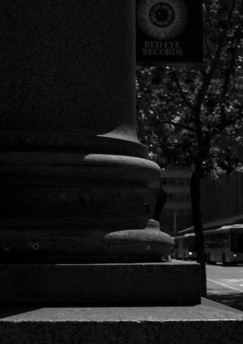 A black and white photo of a classical ionic column in deep shadow with a sign for Red Eye Records in the background.  