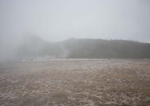 A shrouded and foggy winter field with brown grasses reveals small figures in the distance on a boardwalk, walking through the sulfurous gasses of Yellowstone.