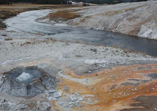 A small geyser bubbles in the foreground, with rusted and sulfuric looking streams pooling off into the river in the distance. 
